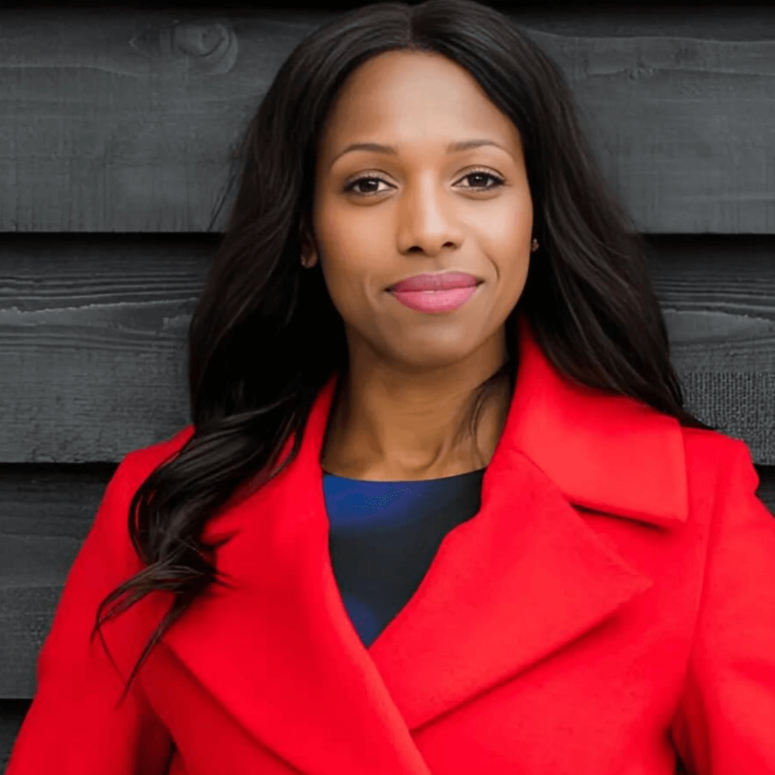 Caroline smiling in front of a dark wooden wall in a red smart coat and long wavy black hair